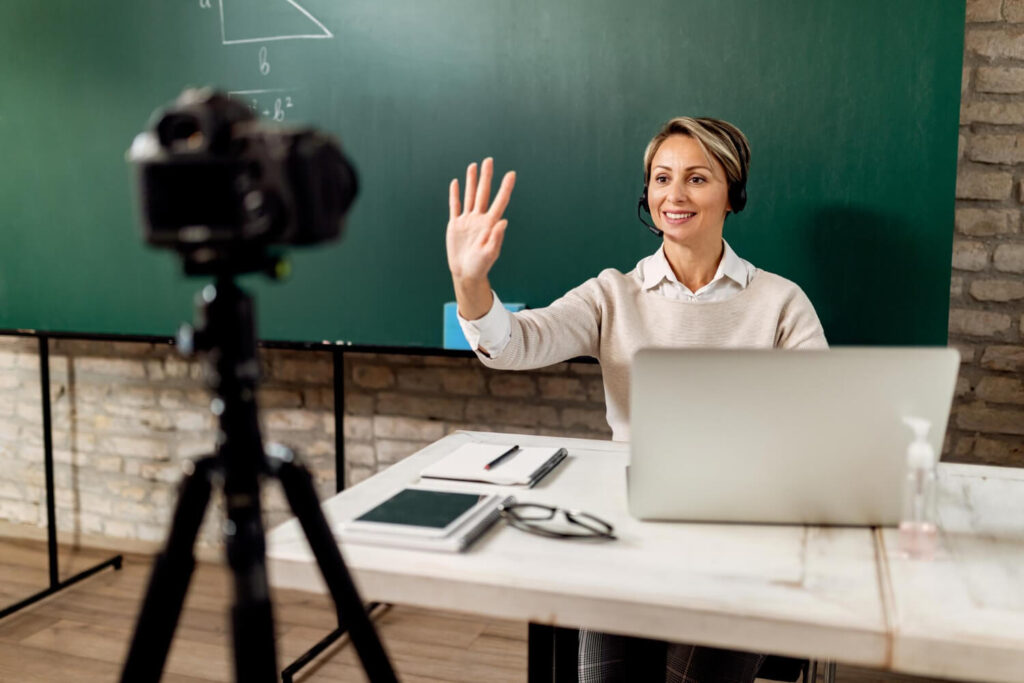 Professora acenando para câmera ao gravar vídeo como aula grátis a ser oferecido como estratégia de marketing educacional.