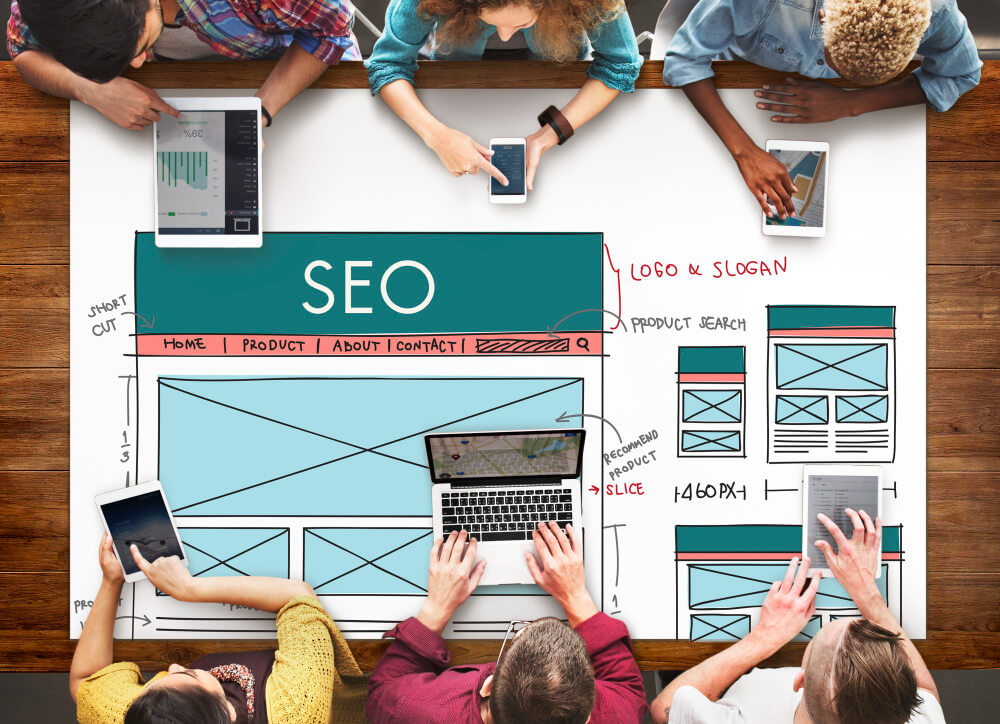 Imagem conceitual de SEO para marketing educacional, com pessoas usando uma mesa de apoio com um esboço de website desenhado em papel mostrando o SEO no topo.